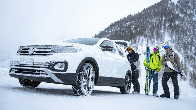 Auto mit Schneeketten im Winterurlaub und Snowboarder im Hintergrund.